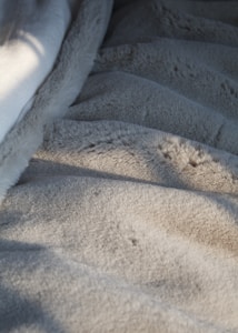A close-up view of a soft, furry grey blanket with subtle shadows cast across its surface, suggesting gentle sunlight coming from one side.