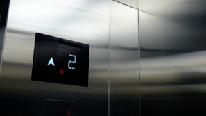 A close-up of an elevator control panel displaying the number two and an upward arrow on a digital screen, surrounded by metallic walls with a reflective surface.