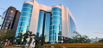 A modern office building with a striking facade of blue-tinted glass windows, interspersed with white panels. The structure is multi-storied with cylindrical sections and is flanked by palm trees and lush greenery. The scene is captured under a clear sky, adding to the building's reflective quality.