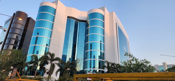 A modern office building with a striking facade of blue-tinted glass windows, interspersed with white panels. The structure is multi-storied with cylindrical sections and is flanked by palm trees and lush greenery. The scene is captured under a clear sky, adding to the building's reflective quality.