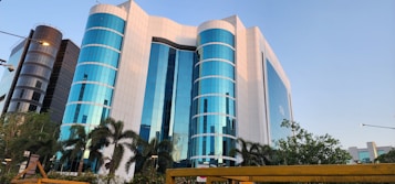A modern office building with a striking facade of blue-tinted glass windows, interspersed with white panels. The structure is multi-storied with cylindrical sections and is flanked by palm trees and lush greenery. The scene is captured under a clear sky, adding to the building's reflective quality.