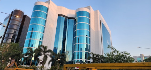 A modern office building with a striking facade of blue-tinted glass windows, interspersed with white panels. The structure is multi-storied with cylindrical sections and is flanked by palm trees and lush greenery. The scene is captured under a clear sky, adding to the building's reflective quality.