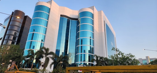 A modern office building with a striking facade of blue-tinted glass windows, interspersed with white panels. The structure is multi-storied with cylindrical sections and is flanked by palm trees and lush greenery. The scene is captured under a clear sky, adding to the building's reflective quality.