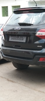 A black SUV is parked, displaying the Ford logo and the model name 'Endeavour' along the rear. The license plate reads 'MP04 EB 1111 ENDEAVOUR'. The vehicle features '4WD' indicating four-wheel drive capability. Part of another vehicle is visible on the left, and there are green metal windows on a wall in the background.