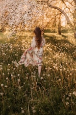 A bright, sunlit scene of a young woman wearing a flowy, colorful dress surrounded by blooming wildflowers.