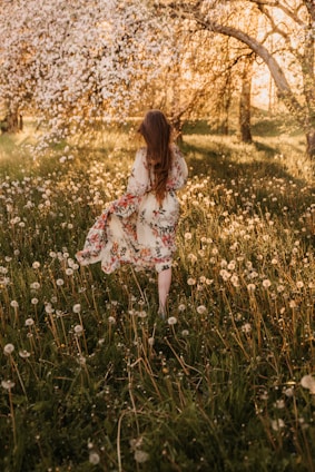 A bright, sunlit scene of a young woman wearing a flowy, colorful dress surrounded by blooming wildflowers.