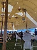 A large tent with a high ceiling is decorated with hanging lanterns and string lights. Inside, several people are seated and standing around wooden tables and chairs. The setting appears to be an event or gathering in an outdoor location, with grass underfoot and a mountainous landscape in the background.