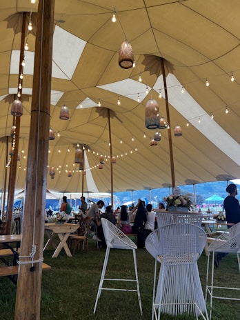 A large tent with a high ceiling is decorated with hanging lanterns and string lights. Inside, several people are seated and standing around wooden tables and chairs. The setting appears to be an event or gathering in an outdoor location, with grass underfoot and a mountainous landscape in the background.