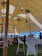 A large tent with a high ceiling is decorated with hanging lanterns and string lights. Inside, several people are seated and standing around wooden tables and chairs. The setting appears to be an event or gathering in an outdoor location, with grass underfoot and a mountainous landscape in the background.