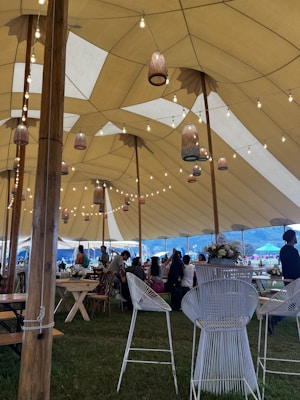 A large tent with a high ceiling is decorated with hanging lanterns and string lights. Inside, several people are seated and standing around wooden tables and chairs. The setting appears to be an event or gathering in an outdoor location, with grass underfoot and a mountainous landscape in the background.