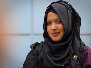 A person wearing a black hijab adorned with a decorative brooch. The background features a softly blurred light blue wall, and the individual is wearing subtle makeup with a warm smile.