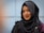 A person wearing a black hijab adorned with a decorative brooch. The background features a softly blurred light blue wall, and the individual is wearing subtle makeup with a warm smile.