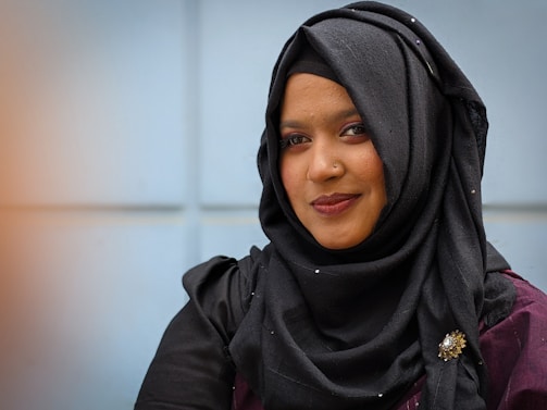 A person wearing a black hijab adorned with a decorative brooch. The background features a softly blurred light blue wall, and the individual is wearing subtle makeup with a warm smile.