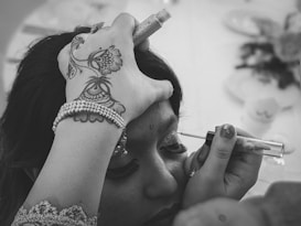 A person is having makeup applied to their eyelids. The hand holding the applicator is adorned with intricate henna designs and a beaded bracelet. The setting seems to be a close-up, with the focus on the makeup process, highlighting the careful attention to detail.