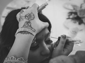 A person is having makeup applied to their eyelids. The hand holding the applicator is adorned with intricate henna designs and a beaded bracelet. The setting seems to be a close-up, with the focus on the makeup process, highlighting the careful attention to detail.