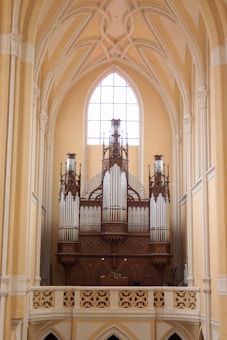 An ornate pipe organ is situated within a grand architectural interior featuring high vaulted ceilings and a large arched window that allows natural light to illuminate the space. The organ is intricately designed with multiple pipes of varying sizes and a decorative wooden frame.