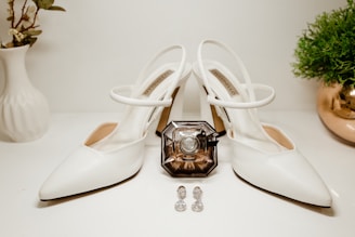 A pair of elegant white high-heeled shoes is positioned centrally, displaying a chic and minimal design. Between the shoes sits an intricately designed perfume bottle with a vintage appearance. Two sparkling earrings with a gemstone-like appearance are placed in front of the perfume. On the left, there is a white vase with some green foliage, and on the right, a small potted plant with lush greenery resides in a reflective golden pot.
