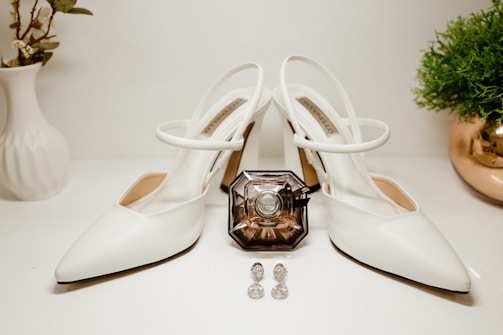 A pair of elegant white high-heeled shoes is positioned centrally, displaying a chic and minimal design. Between the shoes sits an intricately designed perfume bottle with a vintage appearance. Two sparkling earrings with a gemstone-like appearance are placed in front of the perfume. On the left, there is a white vase with some green foliage, and on the right, a small potted plant with lush greenery resides in a reflective golden pot.