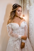A woman dressed in an elaborate white wedding gown adorned with intricate floral lace patterns. She has long, wavy hair styled elegantly with a headpiece. The background includes ornate decorative elements and soft, warm lighting provided by a hanging light bulb.