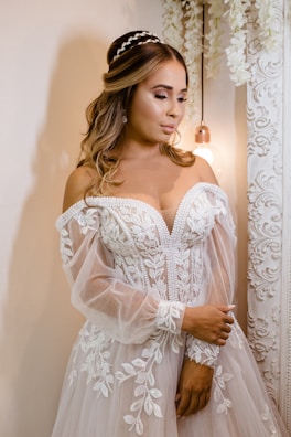 A woman dressed in an elaborate white wedding gown adorned with intricate floral lace patterns. She has long, wavy hair styled elegantly with a headpiece. The background includes ornate decorative elements and soft, warm lighting provided by a hanging light bulb.