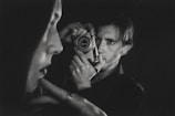A black-and-white photograph captures a person holding a vintage camera up to their face. In the foreground, there is a blurred profile of another person. The composition emphasizes contrast and depth, creating an intimate and introspective atmosphere.