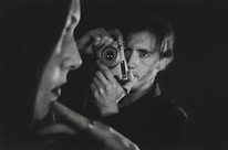 A black-and-white photograph captures a person holding a vintage camera up to their face. In the foreground, there is a blurred profile of another person. The composition emphasizes contrast and depth, creating an intimate and introspective atmosphere.