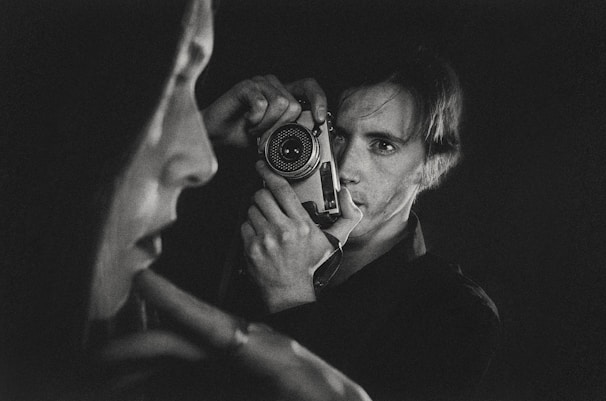 A black-and-white photograph captures a person holding a vintage camera up to their face. In the foreground, there is a blurred profile of another person. The composition emphasizes contrast and depth, creating an intimate and introspective atmosphere.