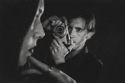 A black-and-white photograph captures a person holding a vintage camera up to their face. In the foreground, there is a blurred profile of another person. The composition emphasizes contrast and depth, creating an intimate and introspective atmosphere.