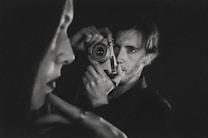 A black-and-white photograph captures a person holding a vintage camera up to their face. In the foreground, there is a blurred profile of another person. The composition emphasizes contrast and depth, creating an intimate and introspective atmosphere.