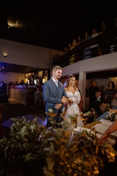 A couple stands at the altar during a wedding ceremony, surrounded by guests in a dimly lit venue. The bride is wearing a white lace dress and a tiara, while the groom is dressed in a blue suit. There are decorations, including greenery and flowers, and an open book is visible on a table nearby.