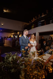 A couple stands at the altar during a wedding ceremony, surrounded by guests in a dimly lit venue. The bride is wearing a white lace dress and a tiara, while the groom is dressed in a blue suit. There are decorations, including greenery and flowers, and an open book is visible on a table nearby.