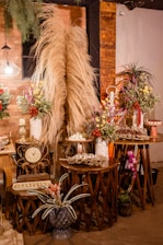 A warm and rustic indoor setting featuring decoratively arranged tables with dry pampas grass, colorful floral arrangements, a vintage clock, and trays of assorted desserts. The backdrop is an exposed brick wall accented with wooden panels and dim lighting from a decorative lamp. Potted plants add a touch of greenery to the scene.
