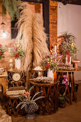 A warm and rustic indoor setting featuring decoratively arranged tables with dry pampas grass, colorful floral arrangements, a vintage clock, and trays of assorted desserts. The backdrop is an exposed brick wall accented with wooden panels and dim lighting from a decorative lamp. Potted plants add a touch of greenery to the scene.