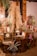 A warm and rustic indoor setting featuring decoratively arranged tables with dry pampas grass, colorful floral arrangements, a vintage clock, and trays of assorted desserts. The backdrop is an exposed brick wall accented with wooden panels and dim lighting from a decorative lamp. Potted plants add a touch of greenery to the scene.