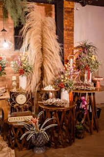 A warm and rustic indoor setting featuring decoratively arranged tables with dry pampas grass, colorful floral arrangements, a vintage clock, and trays of assorted desserts. The backdrop is an exposed brick wall accented with wooden panels and dim lighting from a decorative lamp. Potted plants add a touch of greenery to the scene.
