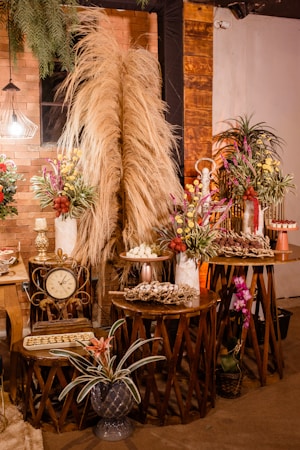A warm and rustic indoor setting featuring decoratively arranged tables with dry pampas grass, colorful floral arrangements, a vintage clock, and trays of assorted desserts. The backdrop is an exposed brick wall accented with wooden panels and dim lighting from a decorative lamp. Potted plants add a touch of greenery to the scene.
