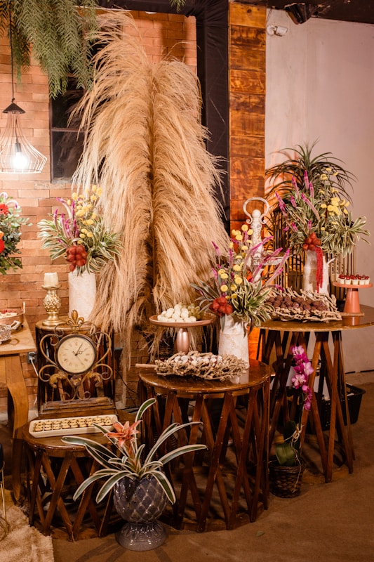 A warm and rustic indoor setting featuring decoratively arranged tables with dry pampas grass, colorful floral arrangements, a vintage clock, and trays of assorted desserts. The backdrop is an exposed brick wall accented with wooden panels and dim lighting from a decorative lamp. Potted plants add a touch of greenery to the scene.