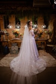 A bride in a long white dress with lace detailing stands in an ornately decorated room. The room features ambient lighting with three hanging lamps, and is adorned with vibrant floral arrangements on a wooden table. There is a backdrop of brick walls and hanging greenery, creating a warm and intimate atmosphere.