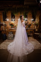 A bride in a long white dress with lace detailing stands in an ornately decorated room. The room features ambient lighting with three hanging lamps, and is adorned with vibrant floral arrangements on a wooden table. There is a backdrop of brick walls and hanging greenery, creating a warm and intimate atmosphere.