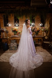A bride in a long white dress with lace detailing stands in an ornately decorated room. The room features ambient lighting with three hanging lamps, and is adorned with vibrant floral arrangements on a wooden table. There is a backdrop of brick walls and hanging greenery, creating a warm and intimate atmosphere.