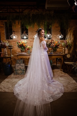 A bride in a long white dress with lace detailing stands in an ornately decorated room. The room features ambient lighting with three hanging lamps, and is adorned with vibrant floral arrangements on a wooden table. There is a backdrop of brick walls and hanging greenery, creating a warm and intimate atmosphere.