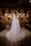 A bride in a long white dress with lace detailing stands in an ornately decorated room. The room features ambient lighting with three hanging lamps, and is adorned with vibrant floral arrangements on a wooden table. There is a backdrop of brick walls and hanging greenery, creating a warm and intimate atmosphere.