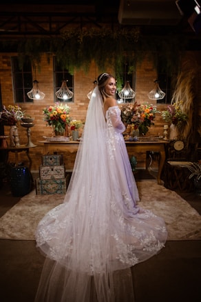 A bride in a long white dress with lace detailing stands in an ornately decorated room. The room features ambient lighting with three hanging lamps, and is adorned with vibrant floral arrangements on a wooden table. There is a backdrop of brick walls and hanging greenery, creating a warm and intimate atmosphere.