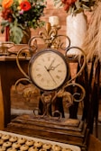 An ornate antique clock with Roman numerals is placed on a rustic wooden table. The background features decorative vases with colorful flowers and dried grasses, adding a warm and elegant atmosphere. The clock has intricate metalwork and a patina finish, suggesting an old-world charm. A tray with small pastries is partially visible in the foreground.
