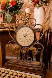 An ornate antique clock with Roman numerals is placed on a rustic wooden table. The background features decorative vases with colorful flowers and dried grasses, adding a warm and elegant atmosphere. The clock has intricate metalwork and a patina finish, suggesting an old-world charm. A tray with small pastries is partially visible in the foreground.