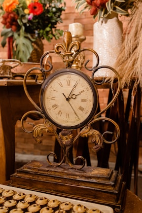 An ornate antique clock with Roman numerals is placed on a rustic wooden table. The background features decorative vases with colorful flowers and dried grasses, adding a warm and elegant atmosphere. The clock has intricate metalwork and a patina finish, suggesting an old-world charm. A tray with small pastries is partially visible in the foreground.