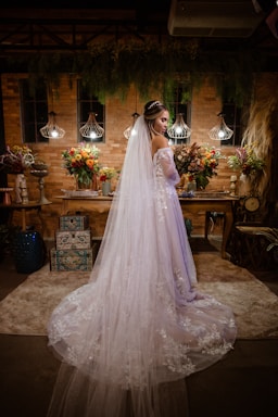 A bride stands in an elegantly decorated room, wearing a long, flowing white gown adorned with lace. Her veil cascades down her back, and she holds a bouquet of flowers. The background features a brick wall with hanging pendant lights and colorful floral arrangements on a wooden table.