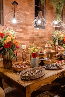 A rustic table setting with an array of desserts and cakes, surrounded by vibrant floral arrangements. The backdrop features a brick wall with decorative light fixtures hanging from above. Various pastries, including trifles and cupcakes, are artfully presented on trays and cake stands. Fresh flowers in shades of red, orange, and yellow add color and warmth to the setting.