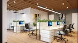 A modern office space with an open-plan layout, featuring multiple white desks arranged in a row and equipped with sleek computers. Comfortable office chairs are positioned around the desks. Green plants are placed on the partitioned tops of the desks, adding a touch of nature to the environment. The room has wooden flooring and a ceiling with exposed wooden beams, along with contemporary lighting fixtures. Artworks hang on the walls, contributing to a creative atmosphere.