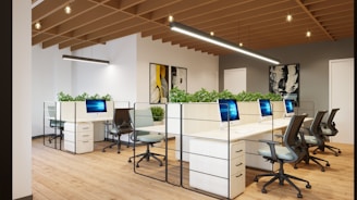 A modern office space with an open-plan layout, featuring multiple white desks arranged in a row and equipped with sleek computers. Comfortable office chairs are positioned around the desks. Green plants are placed on the partitioned tops of the desks, adding a touch of nature to the environment. The room has wooden flooring and a ceiling with exposed wooden beams, along with contemporary lighting fixtures. Artworks hang on the walls, contributing to a creative atmosphere.